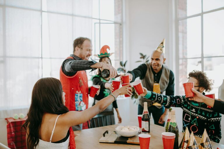 A cheerful group of adults celebrating indoors with holiday attire and festive hats, toasting with red cups.