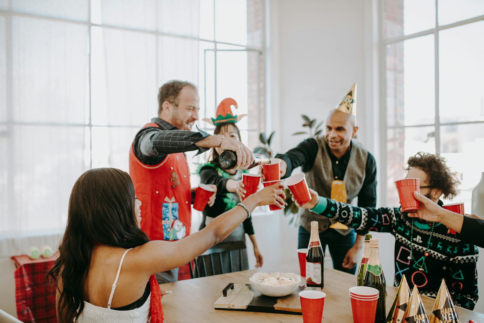 A cheerful group of adults celebrating indoors with holiday attire and festive hats, toasting with red cups.